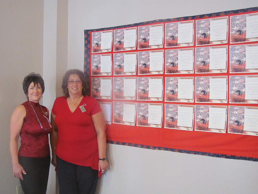 Gayle Raposa (right), the activities director at The Bridge at Cherry Hill, is joined by Sales Director Debra Demar in front of the Veterans Wall of Fame banner Raposa made especially for Monday’s salute.