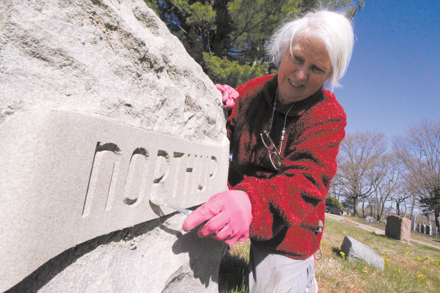 NO TOOTHPASTE: Historic Cemeteries Commission chair Pegee Malcolm uses a tooth brush to clean the lichen that has grown in the lettering of this grave stone. She provided the following outlined of what the commission has planned for May 14 from 1-2:30 pm:  “We will be flagging all the veterans at Brayton ,( there are 260) then we will have a scavenger hunt . Participants will have to find carvings, writings, or symbols on the head stones. These will include items like a headstone taller than you, a headstone with a President’s name, a headstone with your first or last name, a veteran of WW2, a religious symbol on a headstone, a person younger than 10 years old, fruit or flowers carved on a headstone.  There are 25 items to find. Along the way, we will have members of our commission sitting at various graves to explain a bit about that person, such as Thomas Vaill, a Civil war soldier who was on the Ironclad Monitor, Orrin Perry, who was known as Skeleton man in the Barnum and Bailey circus( thank you Kelly Sullivan) Judge Brayton, who was a state Supreme Court justice, . There will also be lawn signs at various headstones to explain the symbolism on them. We are asking folks to use their phones or a camera to take pictures of what they find, but are including a map of the cemetery so in the case of no camera they can circle an area where they find each item. We will be giving prizes to the first 3 people (or groups) that find everything.”