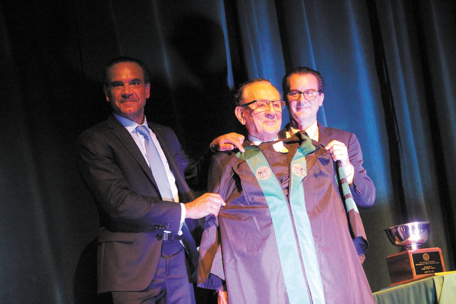 CAUGHT IN WARWICK: Judge Frank Caprio, whose court televised show “Caught in Providence” has earned him national attention for his compassion and bench dialogue with offenders, was presented a graduation robe (Fr. Robert Marciano, Hendricken president, suggested he wear it for the show) after being made an honorary Hendricken graduate.  Helping Judge Caprio with the robe are his sons Frank and David, both Hendricken graduates.